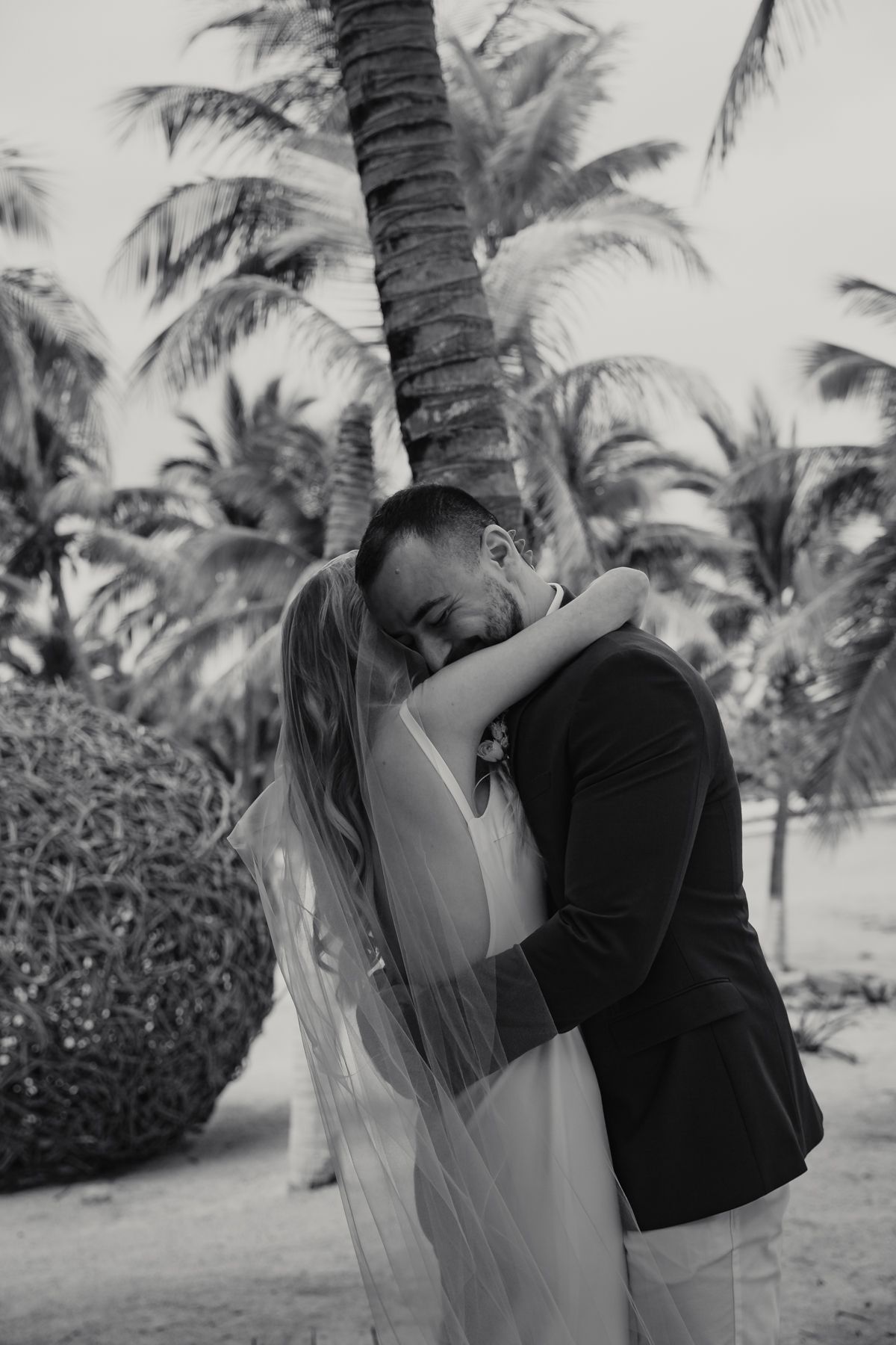 Black and white photo of bride and groom sharing tender embrace near palm trees on tropical beach wedding day.