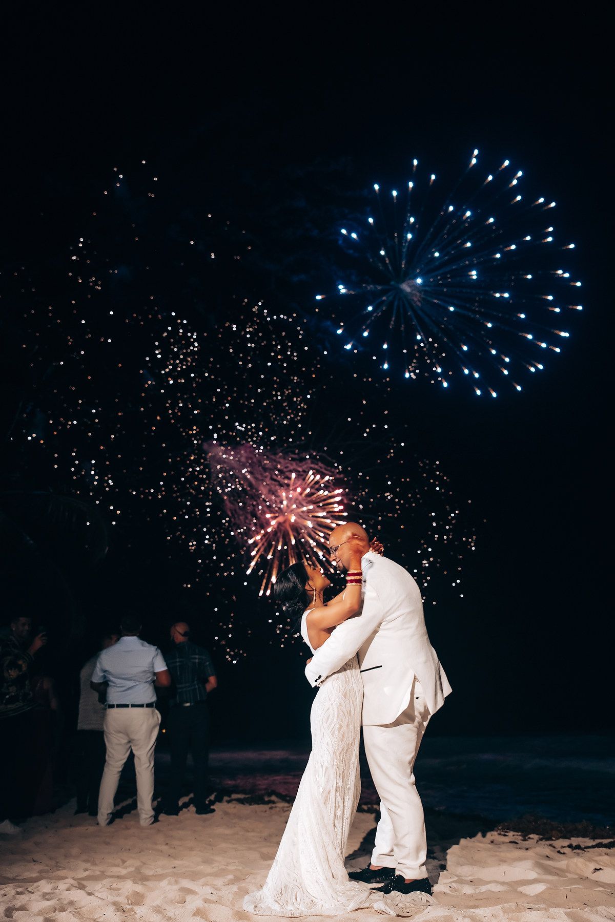 A couple in white attire shares a romantic kiss on the beach while fireworks burst in the dark sky above.