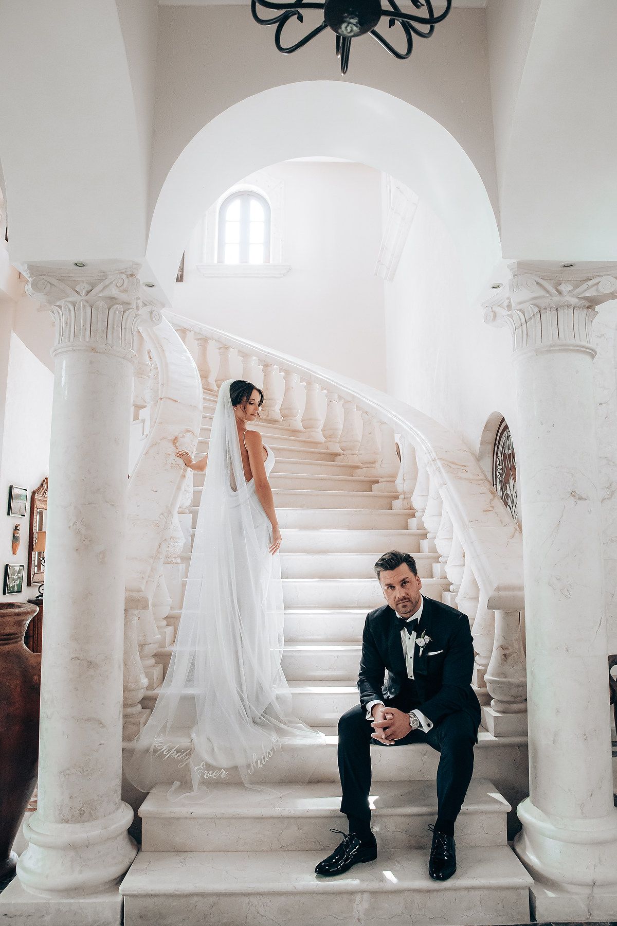 Elegant wedding photo series showing a person in black suit sitting on white marble steps in a classic architectural setting.