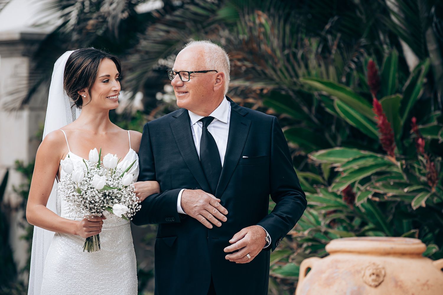 A bride in a white wedding dress holds a bouquet while walking with a gentleman in a dark suit outdoors.