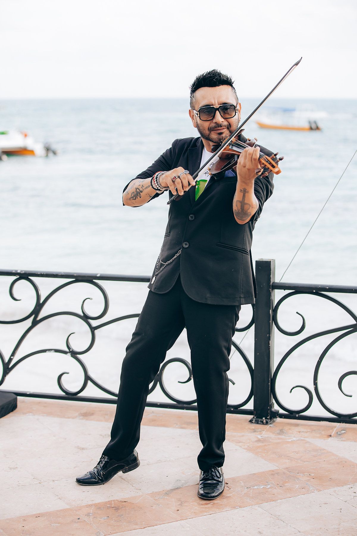 Musician in black suit plays violin on oceanfront terrace with ornate railing and boats visible in background.
