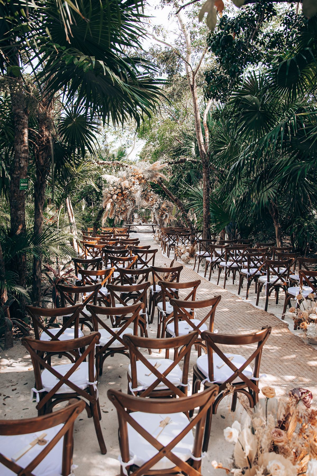 Tropical garden wedding setup with wooden cross-back chairs arranged in neat rows under swaying palm trees.