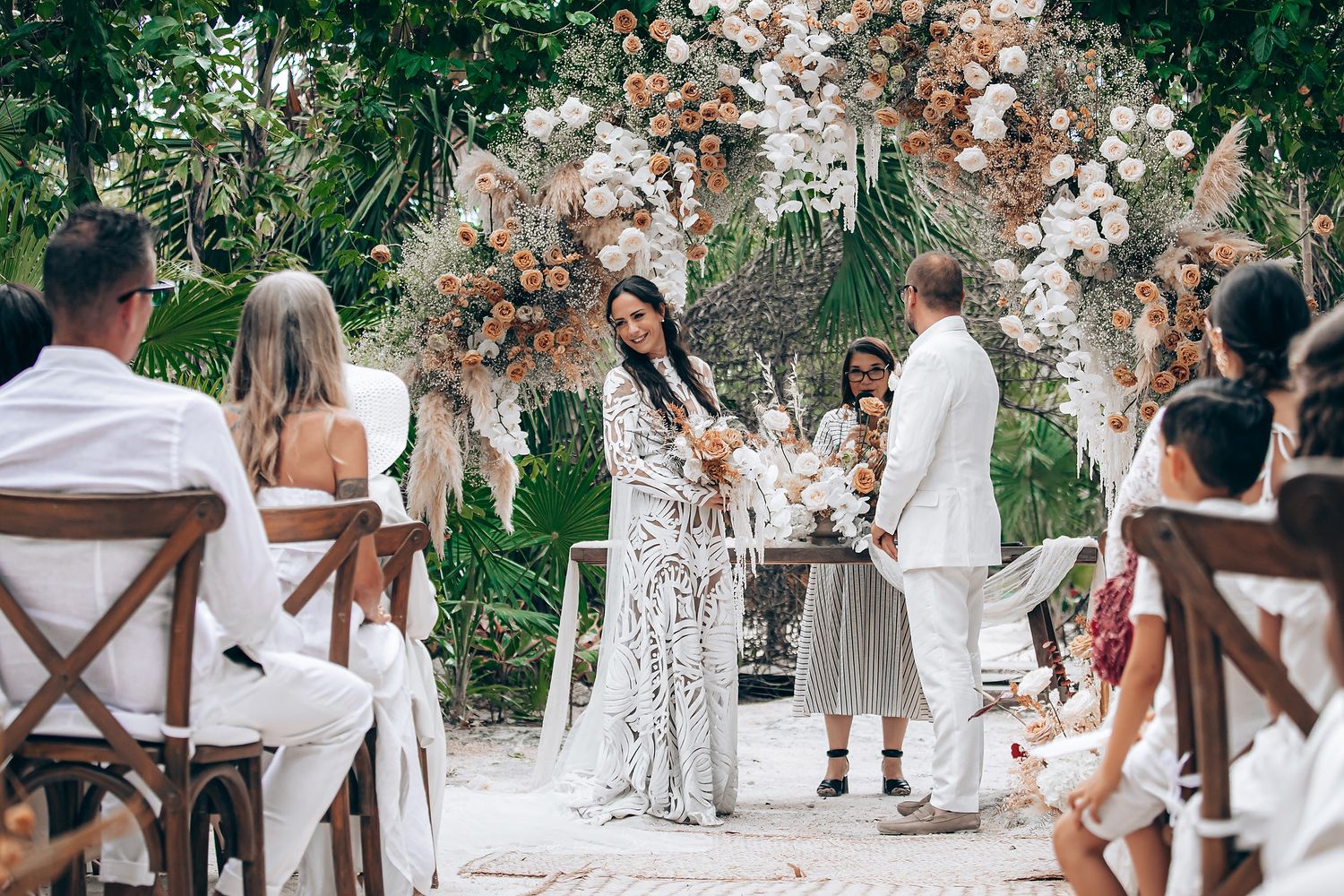 Outdoor wedding ceremony with guests seated on wooden chairs and floral arch decorations in a garden setting.