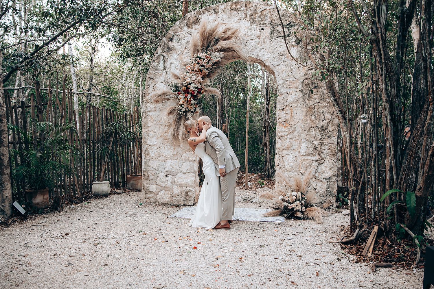 A romantic stone archway decorated with dried flowers stands in a wooded path during a wedding ceremony.