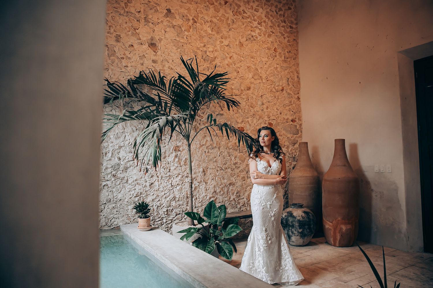 A person in a white lace wedding dress stands against a textured wall with tropical plants and terracotta pots.