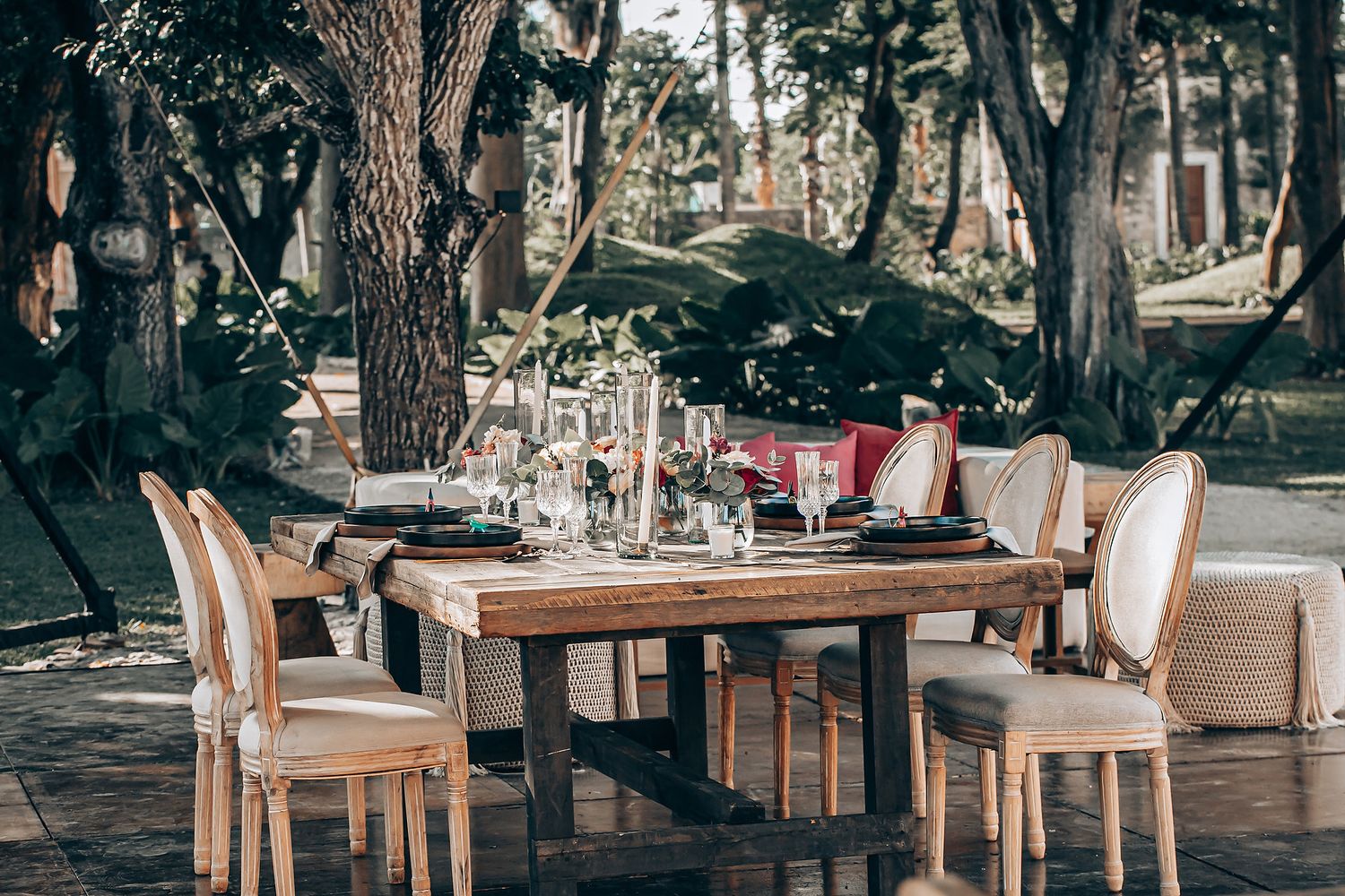 Elegant outdoor wedding reception setup with wooden tables and chairs arranged under tall trees in a forest setting.