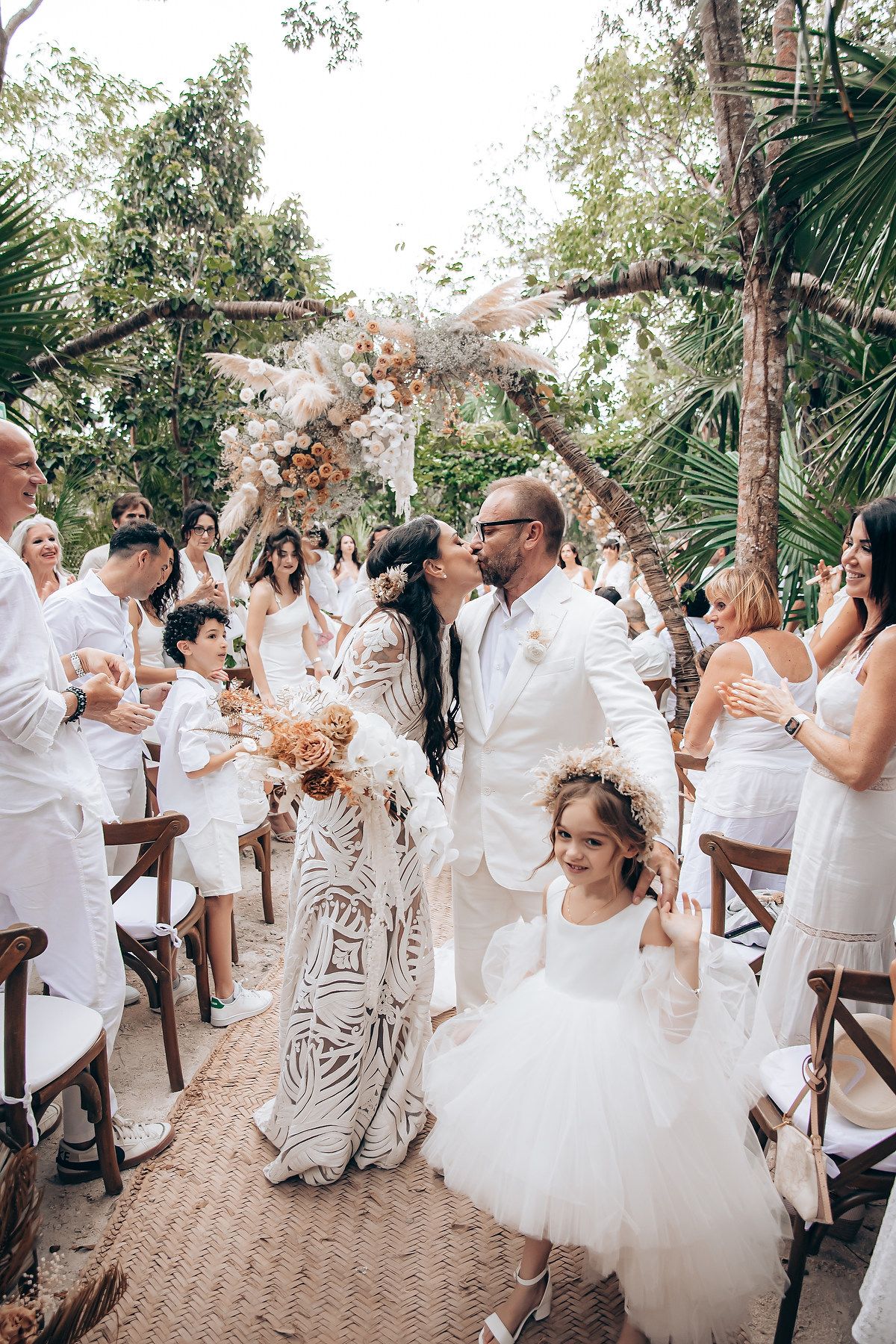 Wedding guests in white attire dance and celebrate at an outdoor tropical venue.