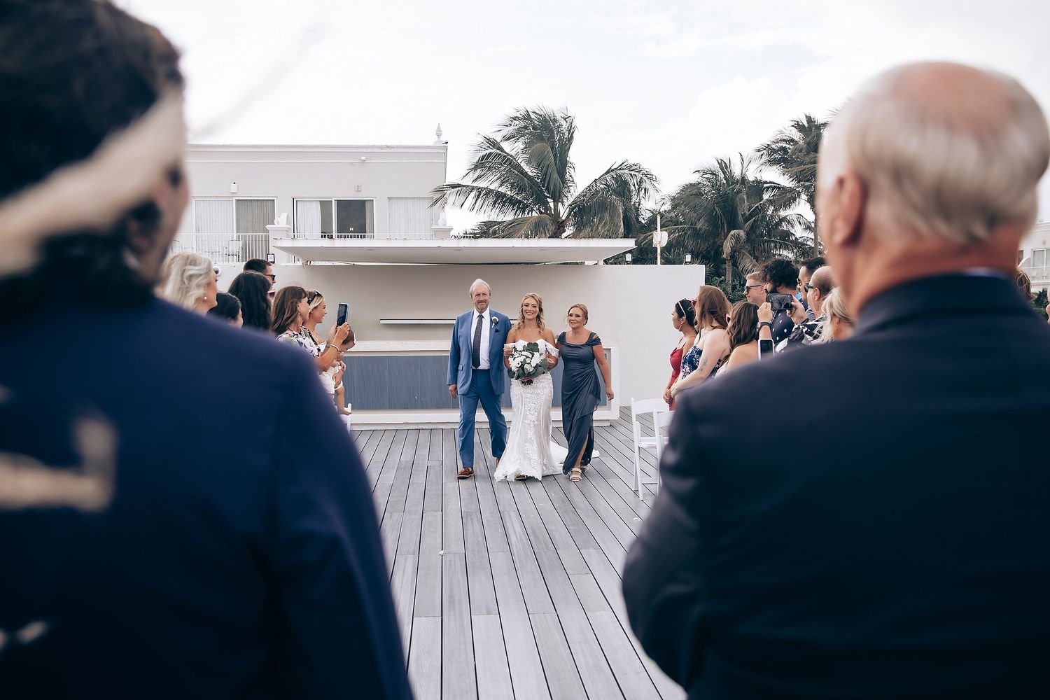 Wedding guests seated on white chairs at an outdoor ceremony with palm trees swaying in the wind.