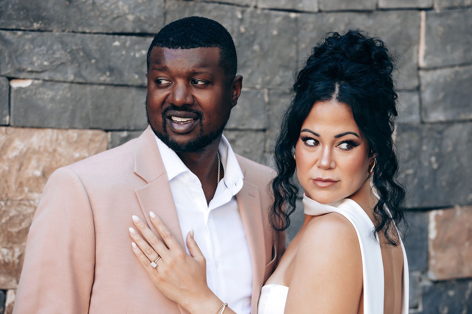 A romantic wedding couple embraces while standing against a gray stone wall backdrop.