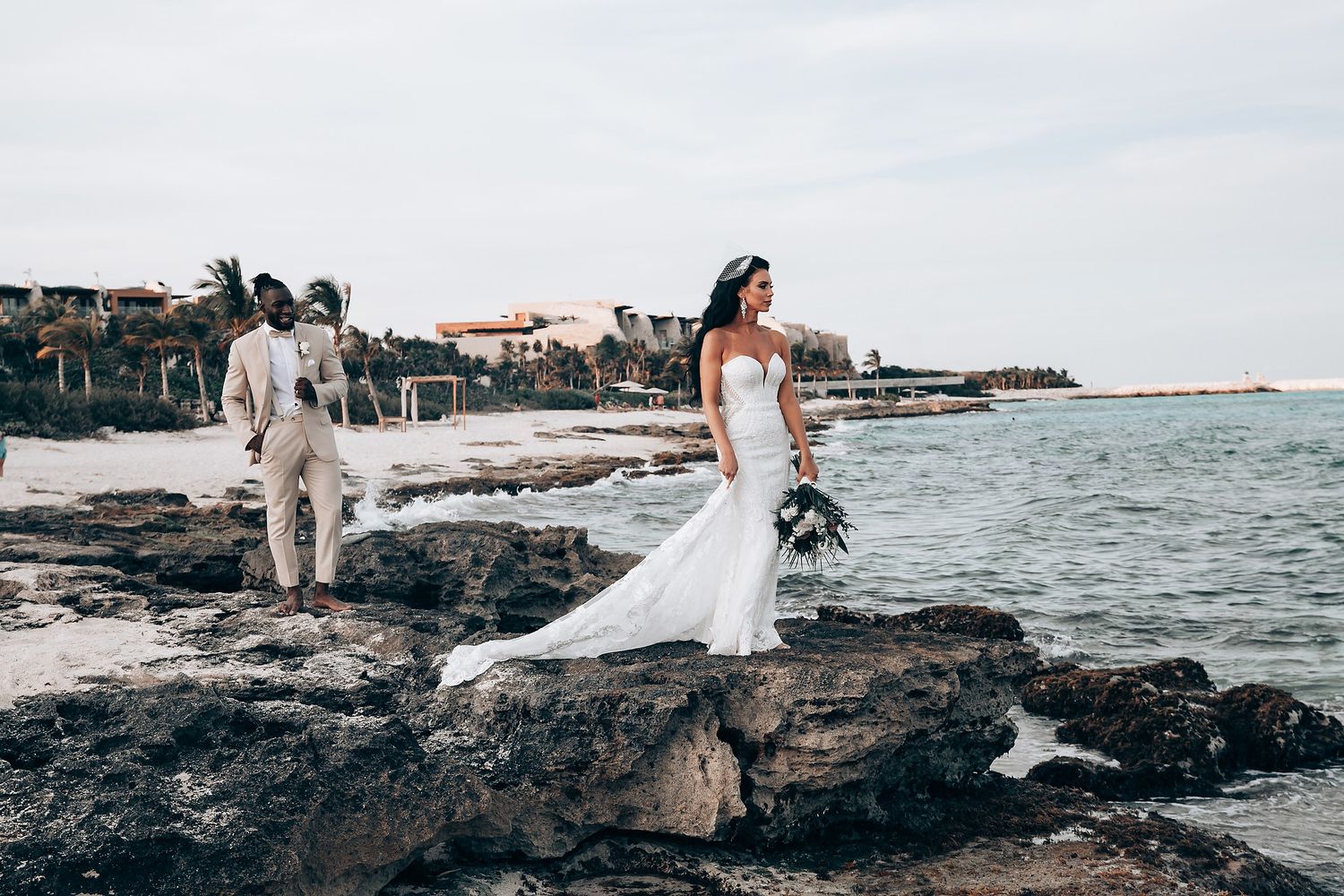Bride in flowing white dress poses on rocky coastal shoreline during wedding photoshoot.