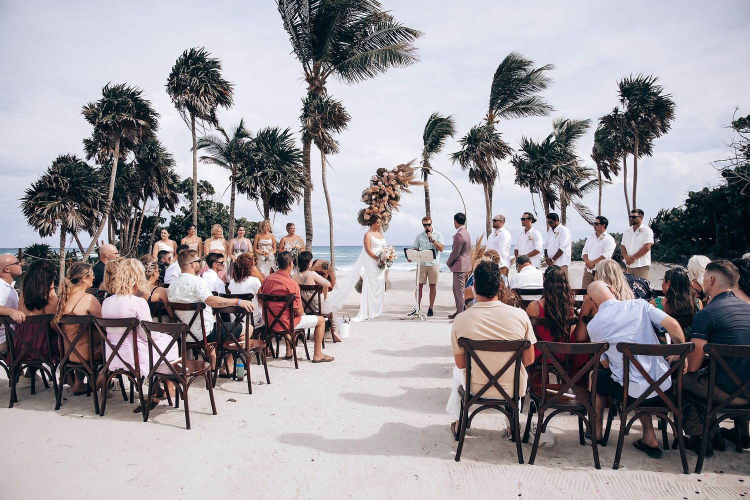 Wedding reception setup on a beach with wooden chairs and palm trees against an overcast sky.