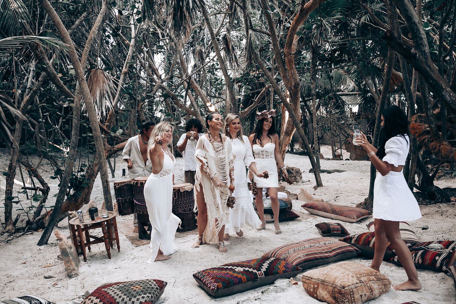 A beach gathering with people lounging on colorful rugs under tropical trees.
