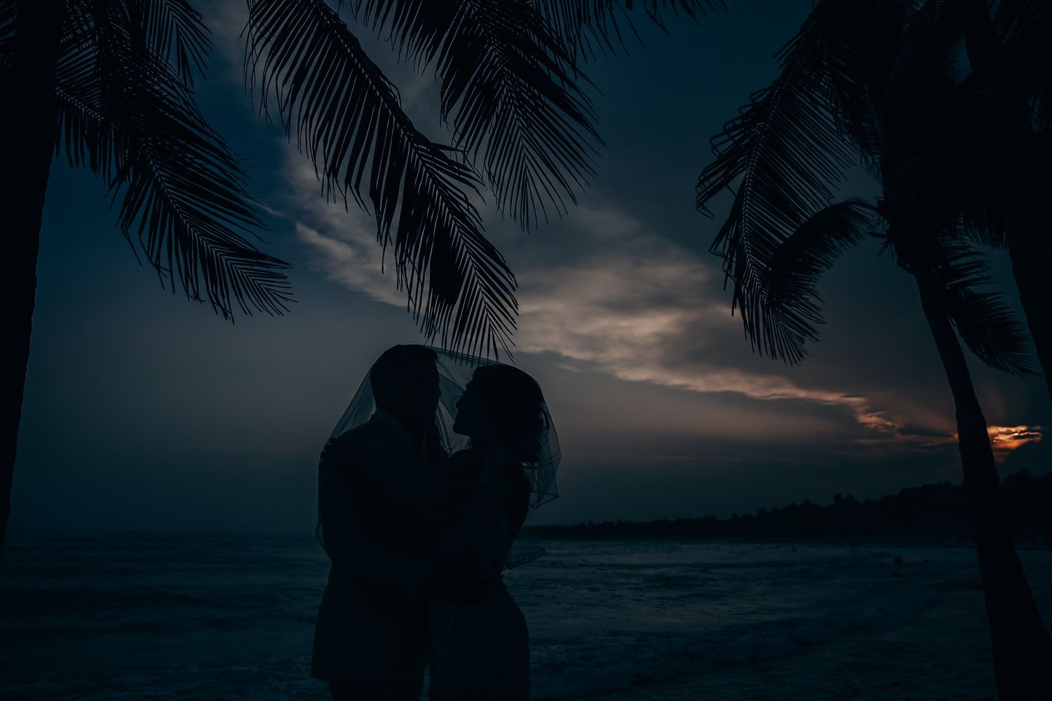 Silhouetted palm trees frame a dramatic sunset over a tropical beach with orange and purple sky.