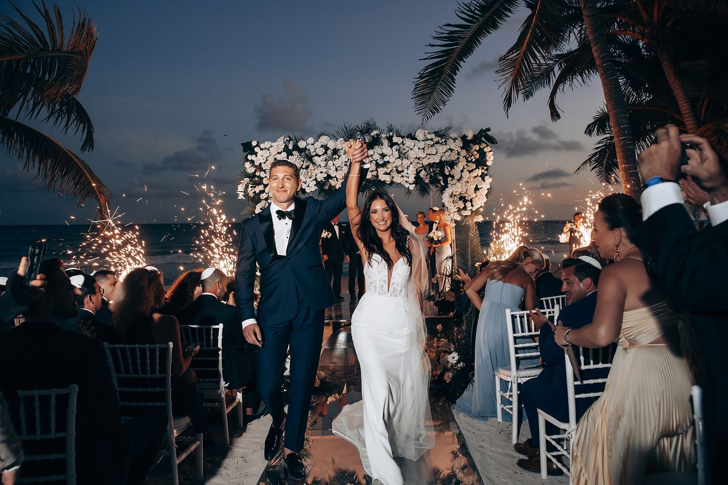 A wedding celebration at sunset with palm trees and guests dancing on an outdoor terrace overlooking the ocean.