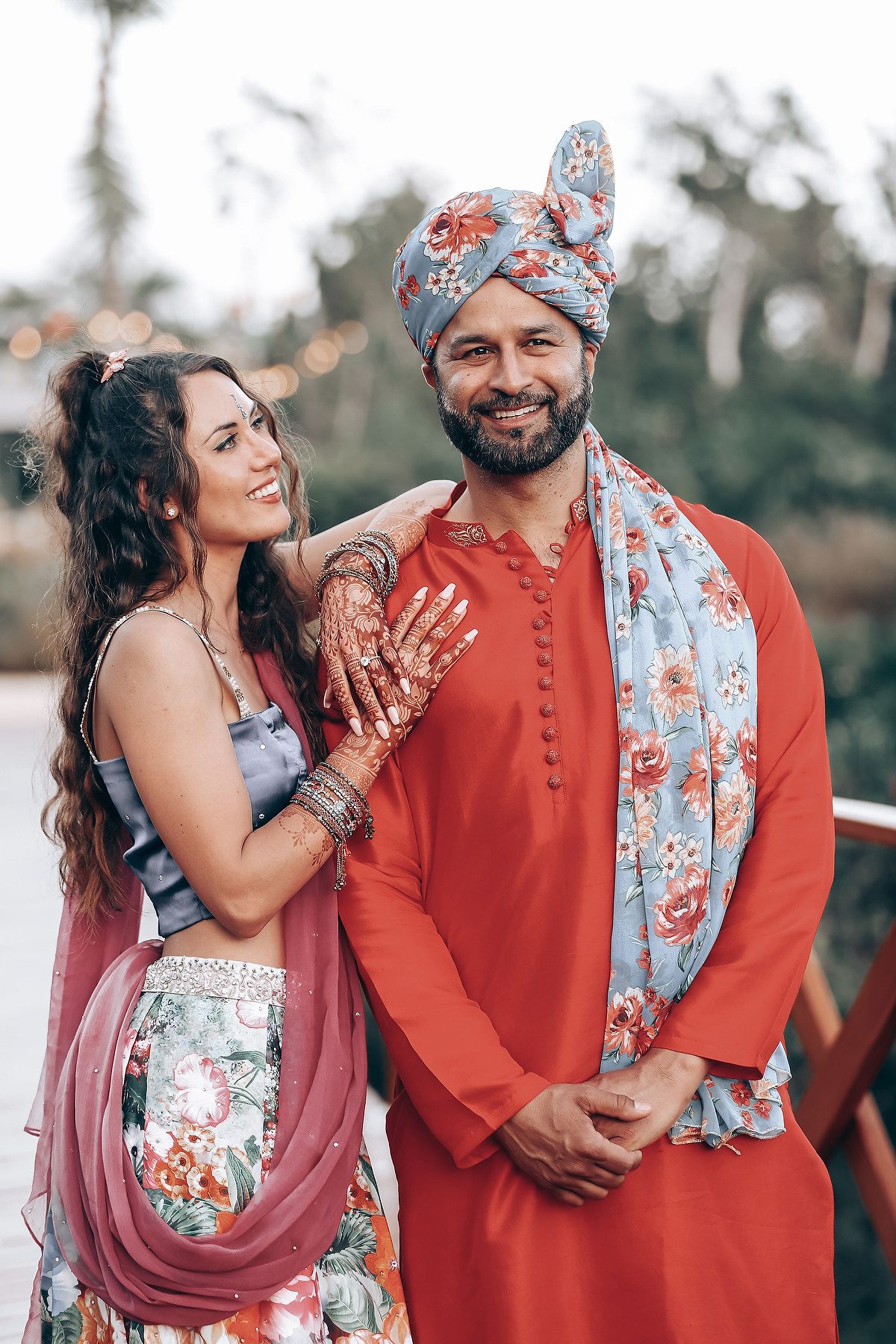 A couple poses together in coordinated Indian outfits featuring orange and floral patterns during a photoshoot.
