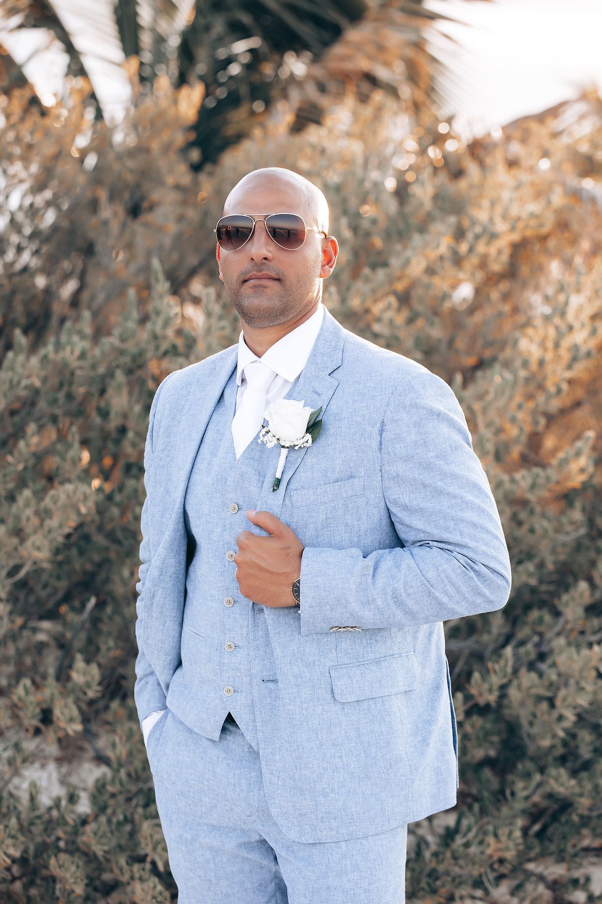 Groom in light blue suit posing against autumn foliage background.