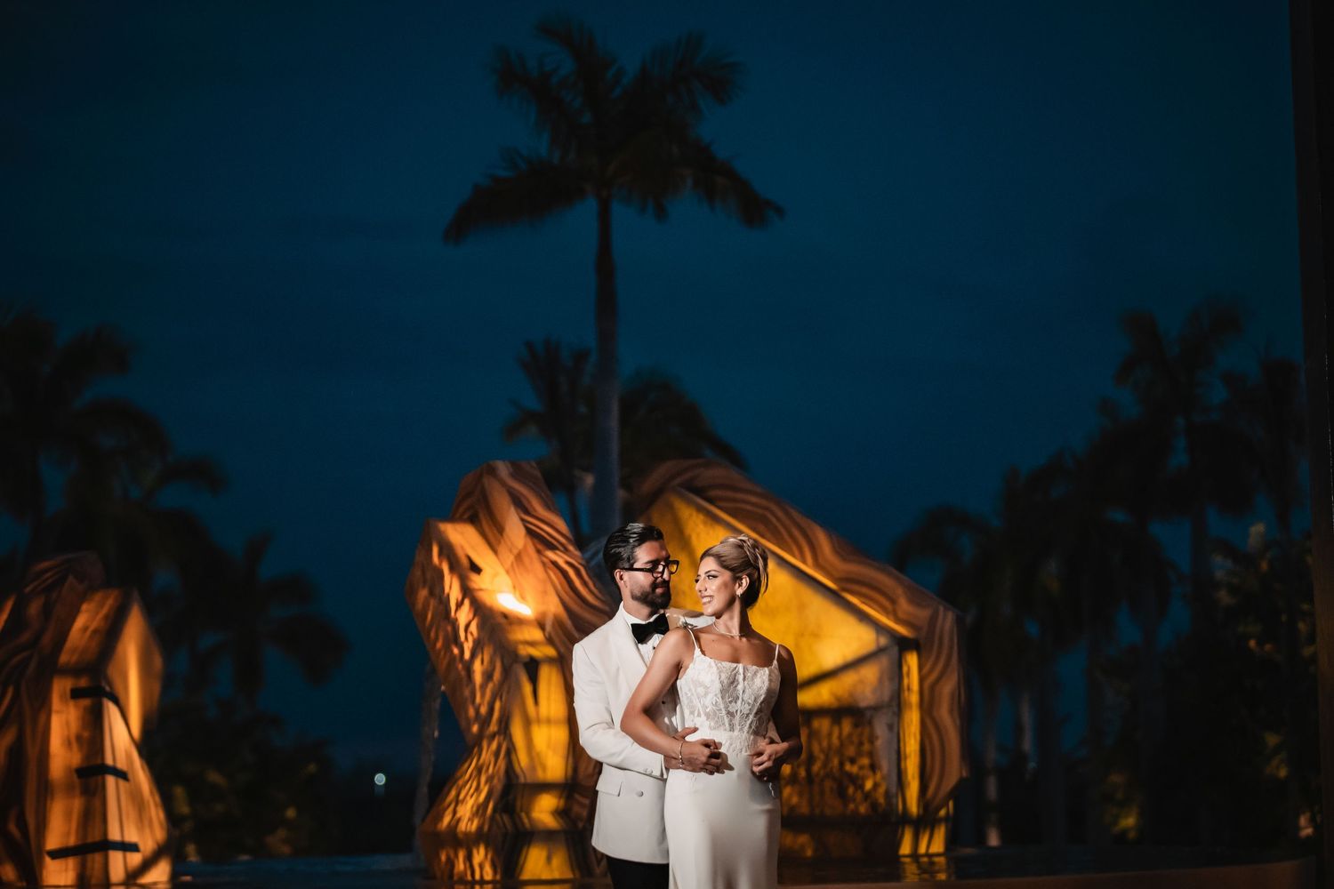 Evening wedding portrait with palm trees and dramatic lighting creating silhouettes.