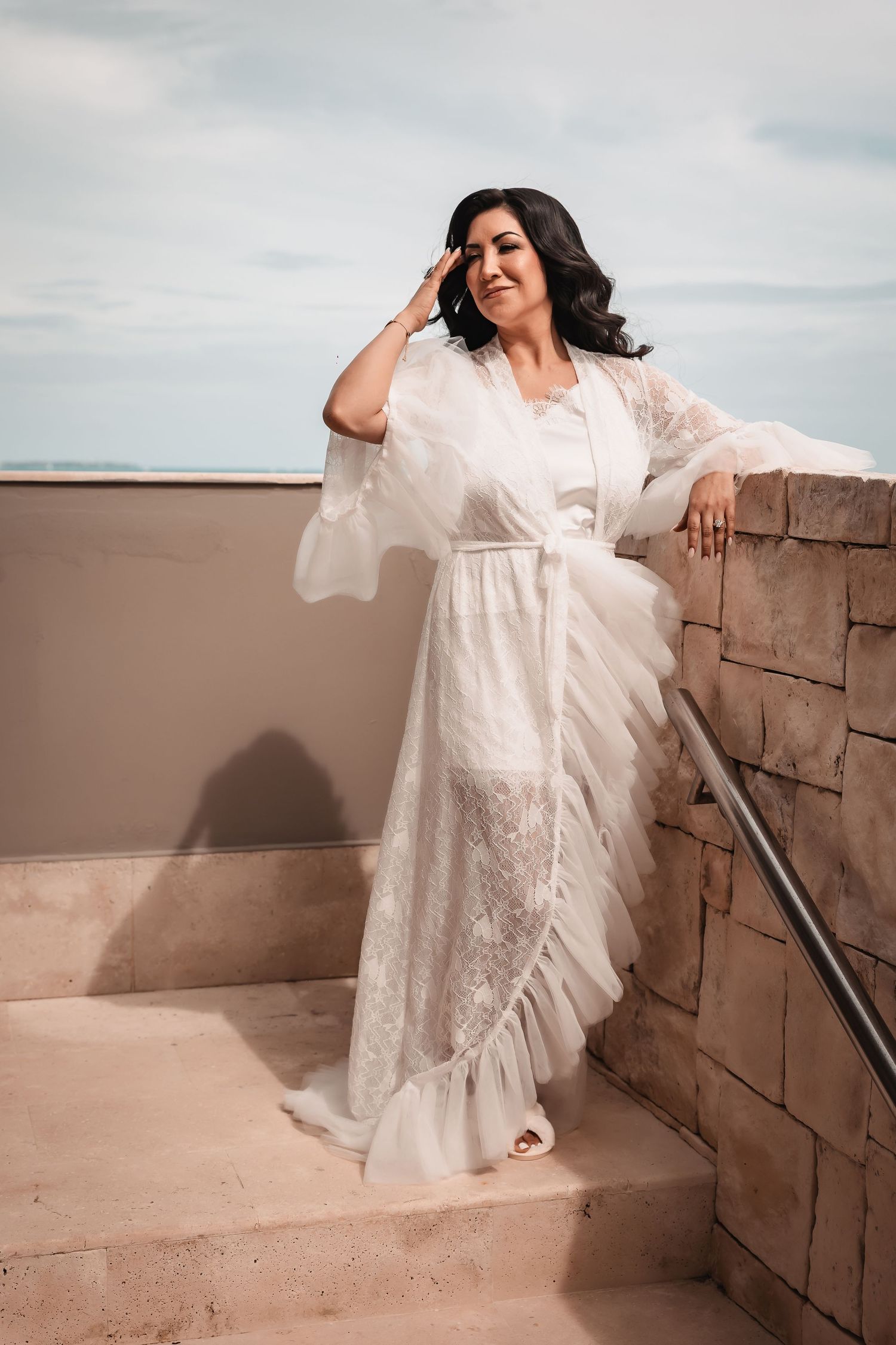 Someone in a flowing white lace robe poses casually on outdoor stone steps by the sea.