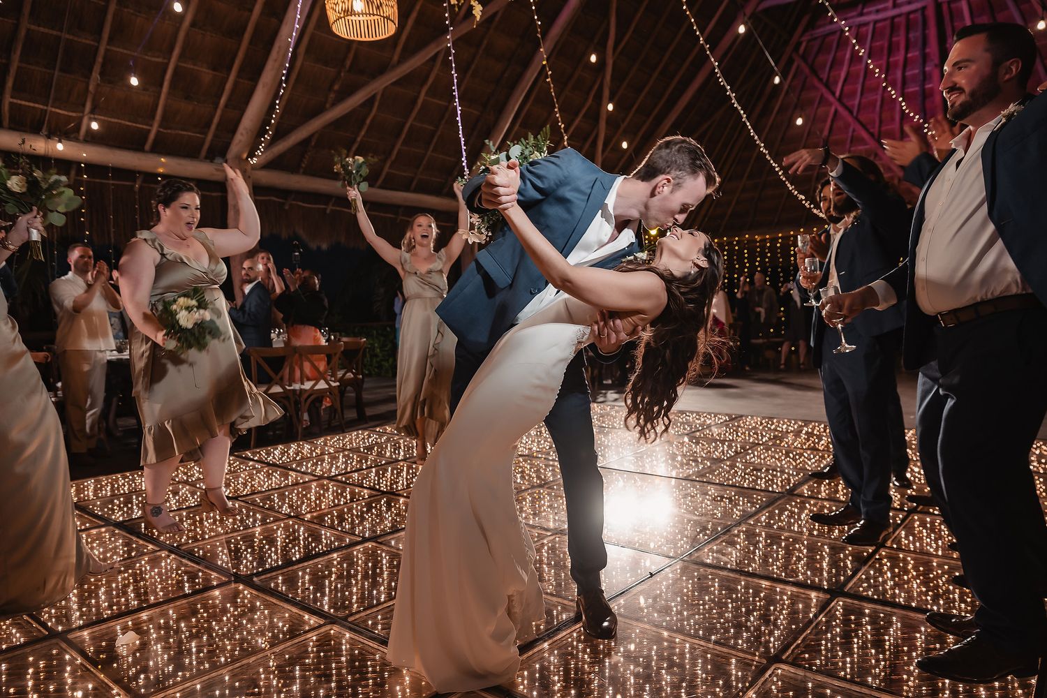 Wedding guests dancing and celebrating on illuminated dance floor in rustic barn setting.