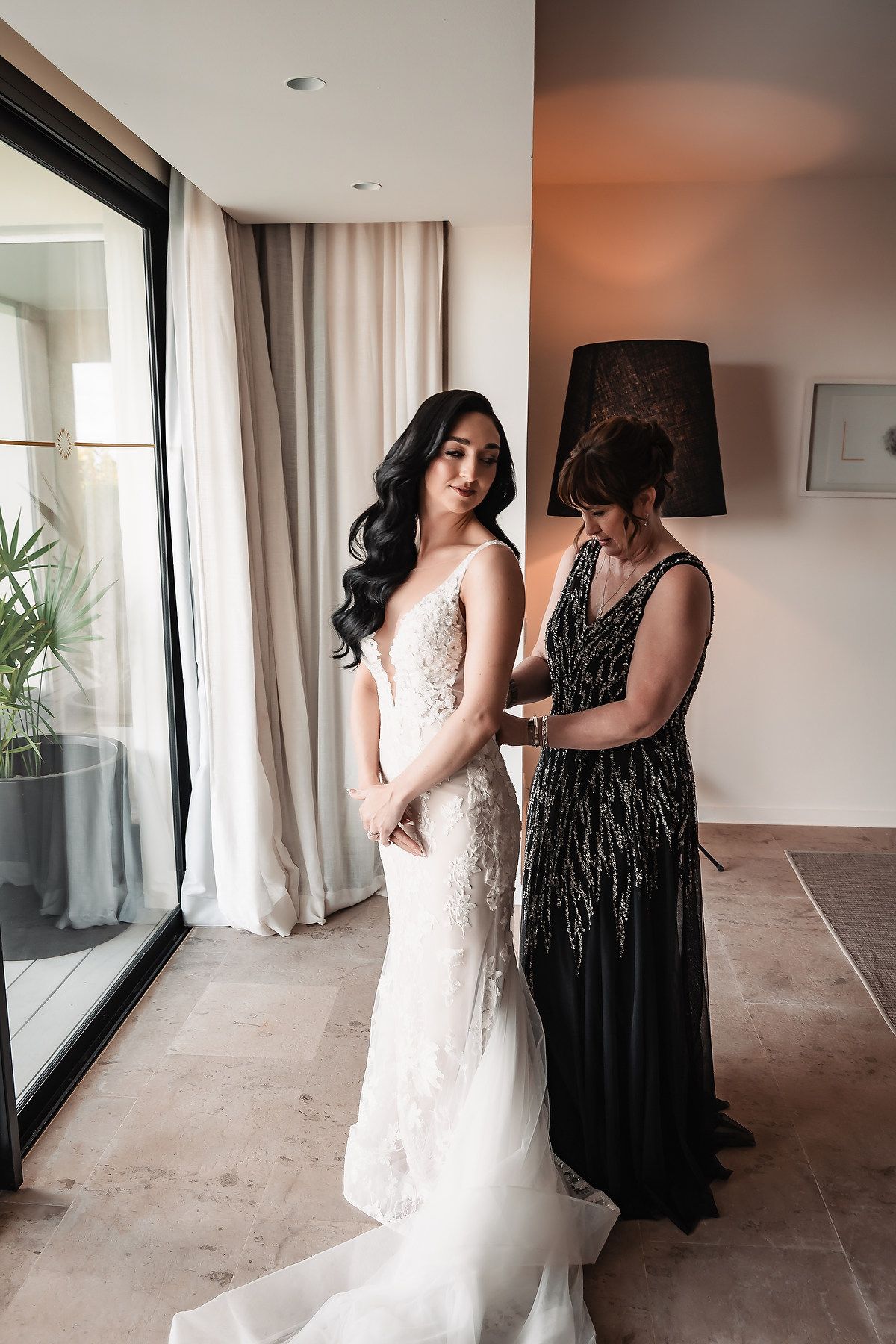 A bride in a white lace wedding dress gets final adjustments in a modern room with floor-to-ceiling windows.