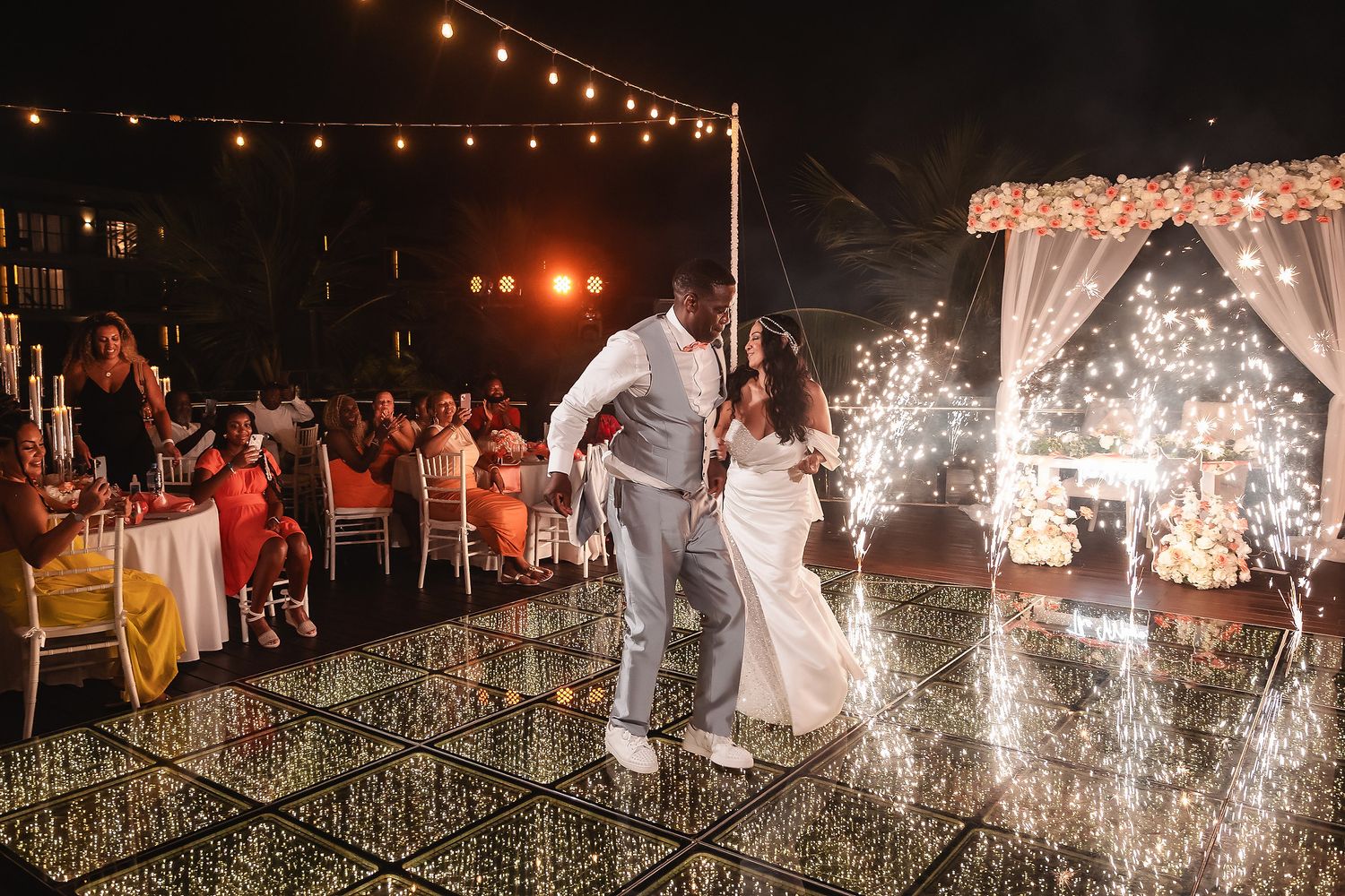 A couple shares their first dance under string lights with sparklers and floral decorations.