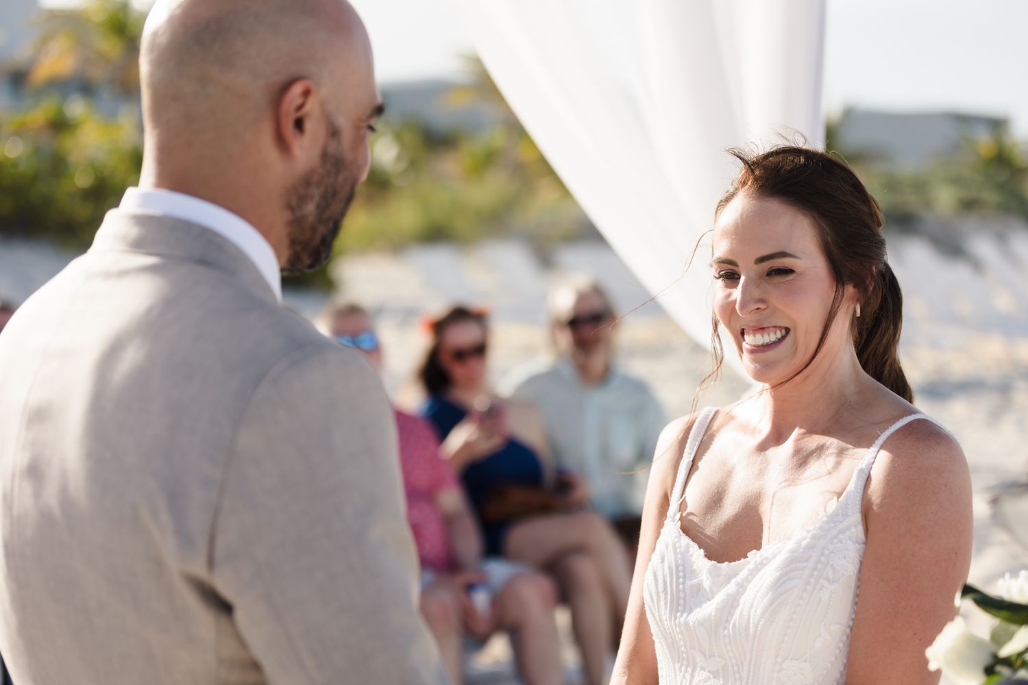 Bride smiling joyfully at groom during outdoor beach wedding ceremony with guests seated in background.