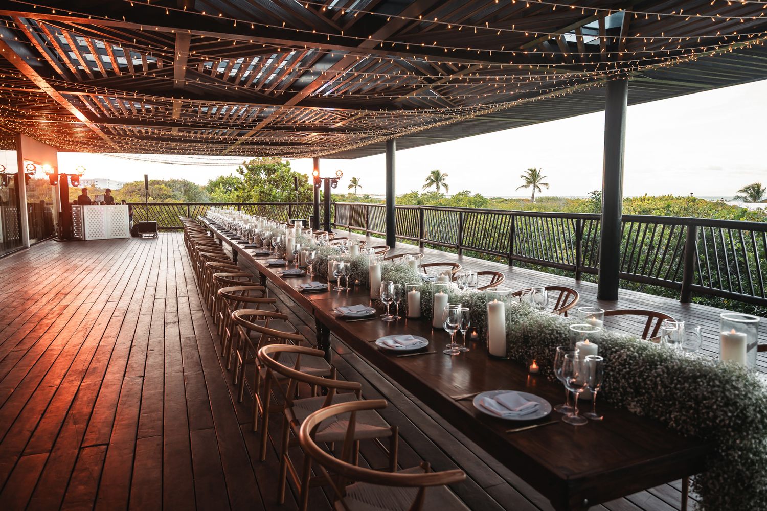 Elegant outdoor wedding reception on a wooden deck at sunset with long dining tables, candles, and string lights.