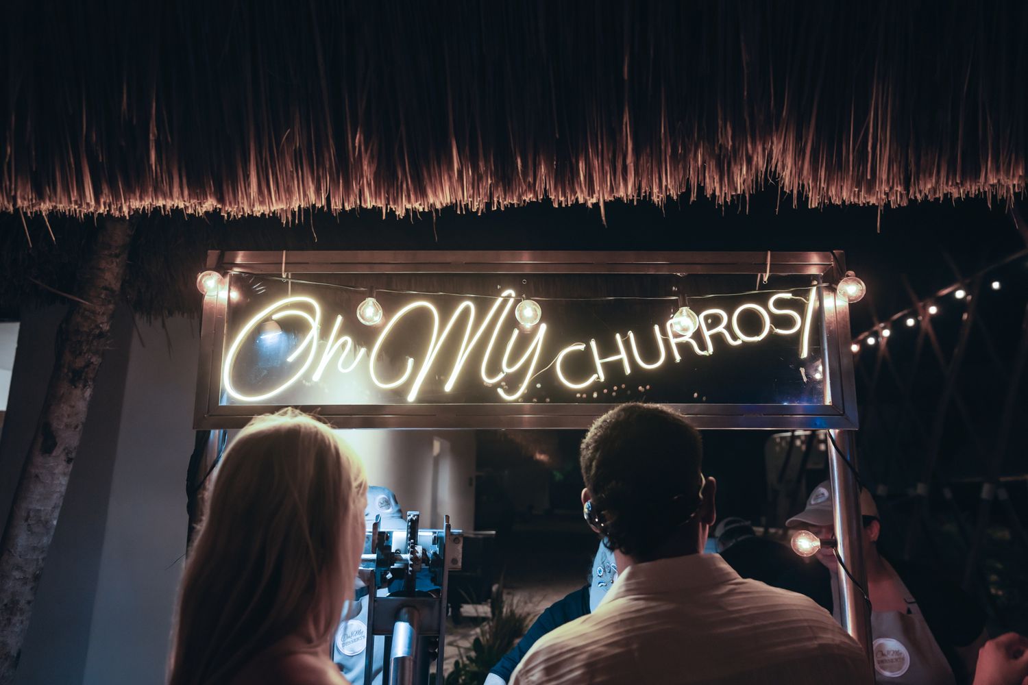Neon sign reading Oh My Churros at a thatched roof food stall with customers waiting.
