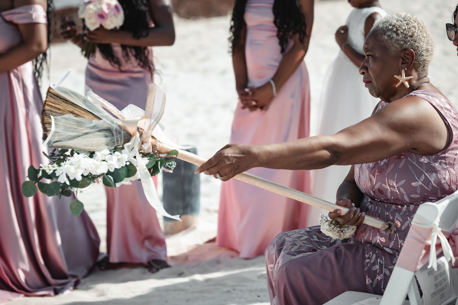 Elderly woman in floral dress releasing a white dove at an outdoor beach wedding ceremony with bridesmaids in pink gowns.
