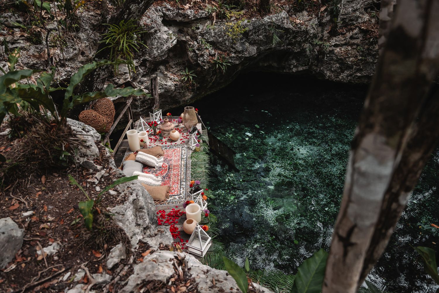 Aerial view of a colorful picnic setup on rocky ledge beside a turquoise cenote in a lush cave.