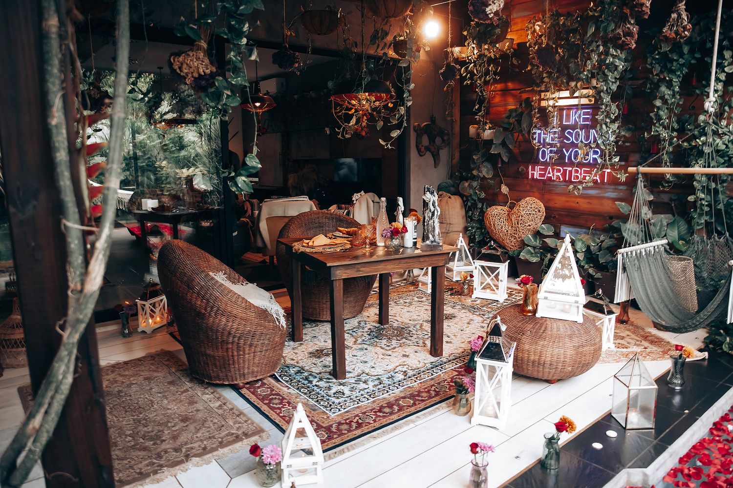 Bohemian indoor lounge with wicker chairs, patterned rugs, lanterns, and neon sign surrounded by lush greenery.