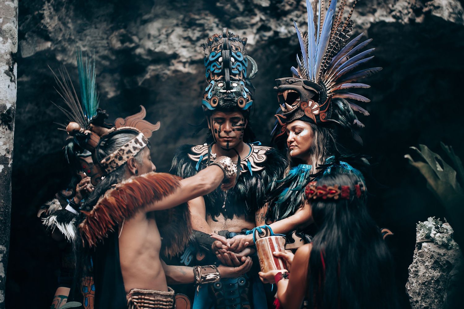 Mayan performers in ornate feathered headdresses and traditional costumes participate in an ancient ceremonial ritual in a cave.