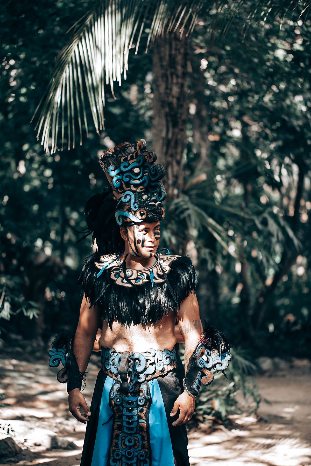 Man in elaborate blue Mayan warrior costume with decorative headdress standing in tropical jungle.