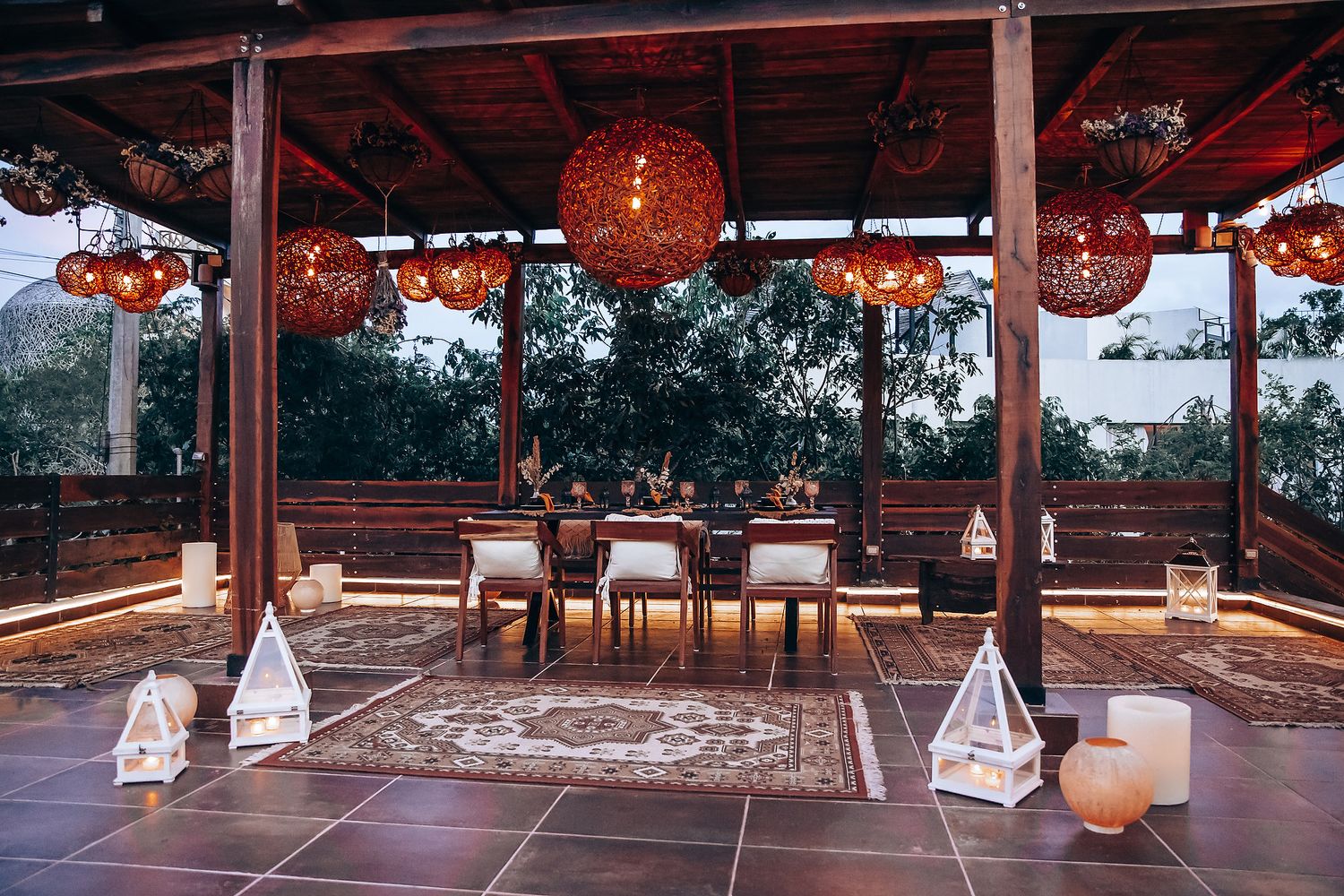 Bohemian outdoor dining pavilion with woven rattan pendant lights, Persian rugs, and pyramid lanterns at dusk.