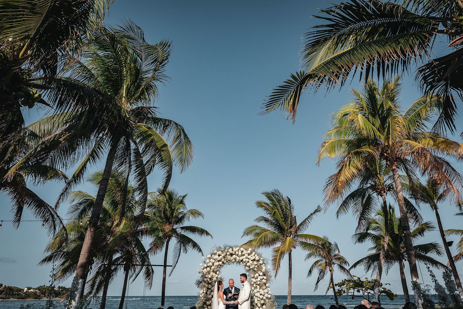 Couple exchanges vows under floral arch surrounded by tall palm trees on a tropical beach wedding.