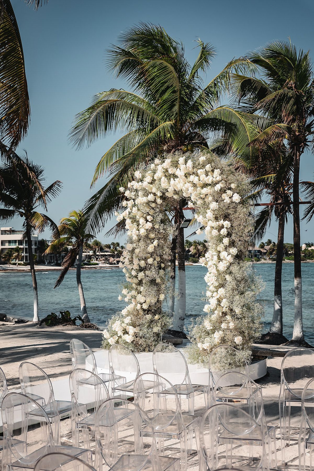 Elegant beachside wedding ceremony setup with white floral arch, ghost chairs, palm trees, and ocean views under blue sky.