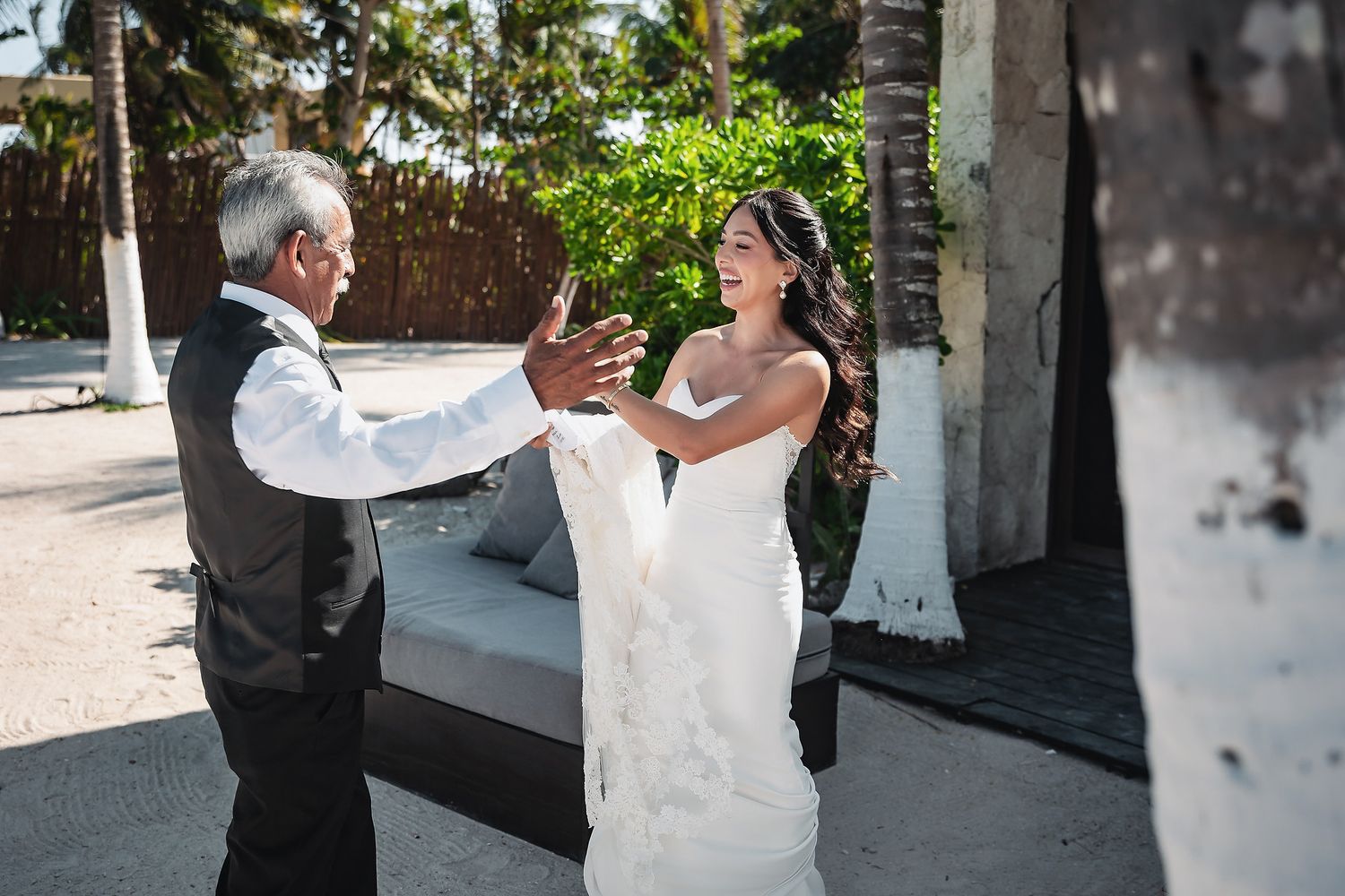 Bride in white gown shares emotional first look moment with father on sandy beach near tropical palm trees.