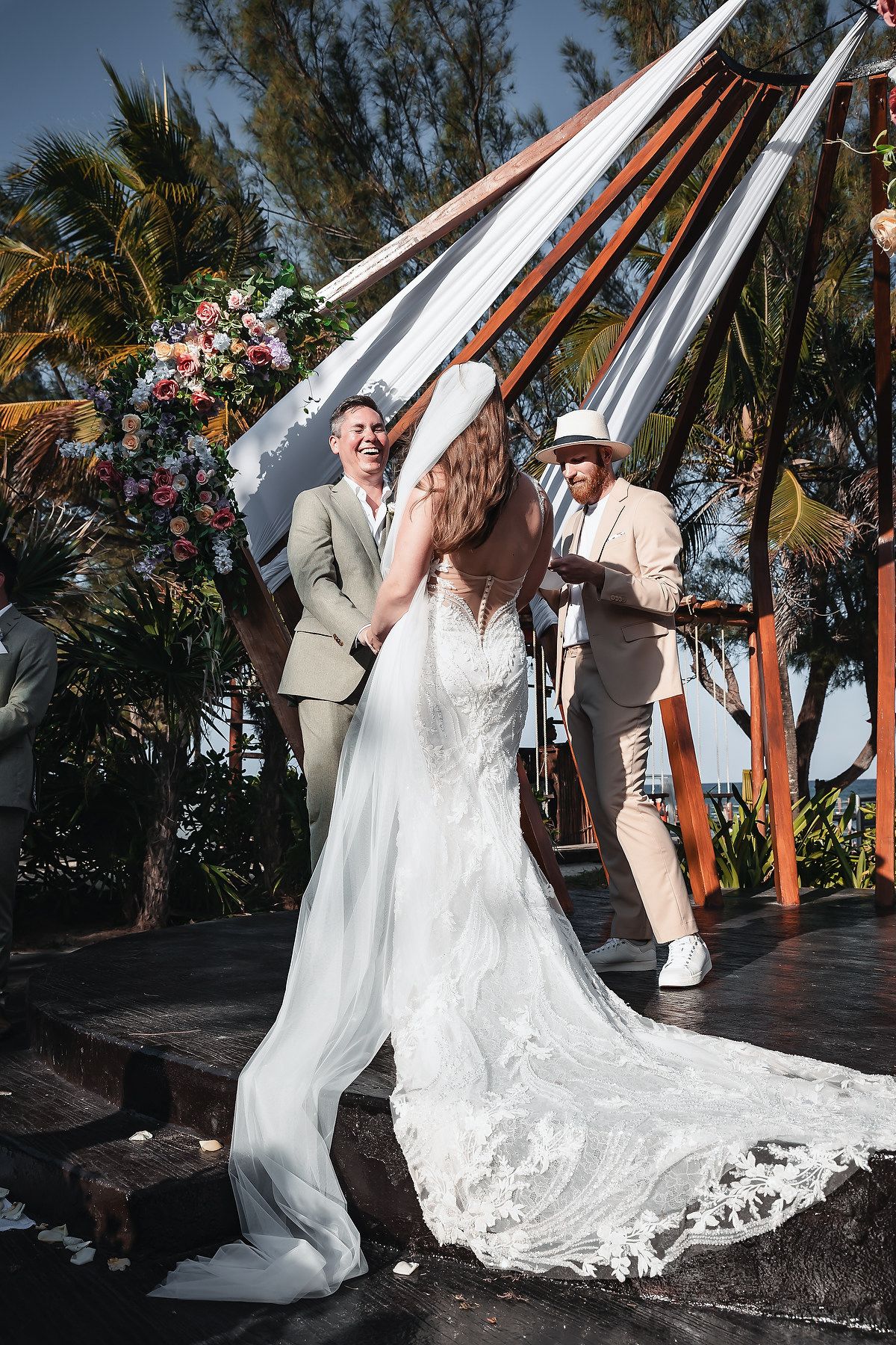 Bride and groom exchange vows at tropical outdoor wedding ceremony with floral arch and palm trees.