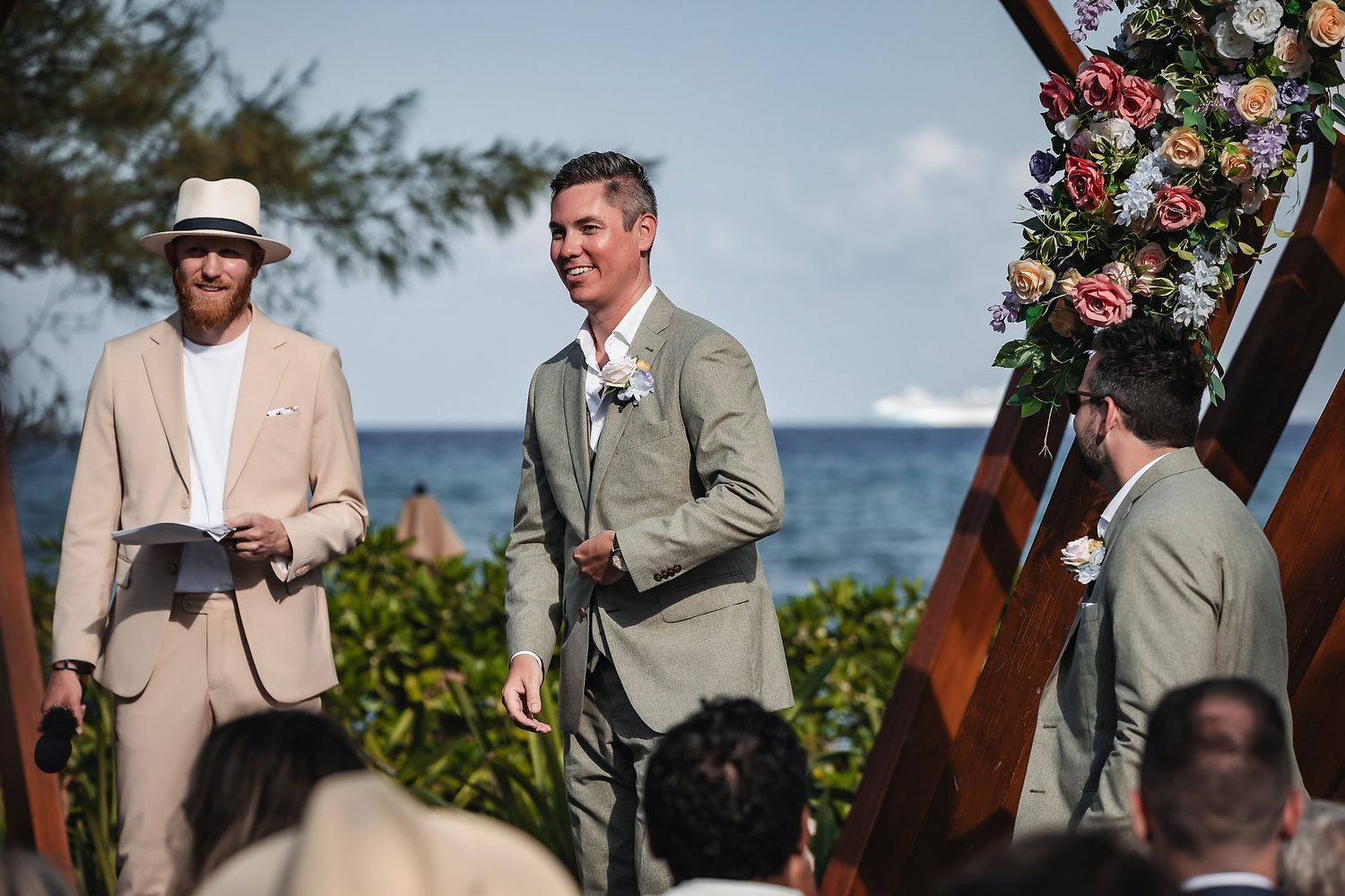 Groom smiling at outdoor beach wedding ceremony with floral arch, officiant in panama hat, and ocean view backdrop.