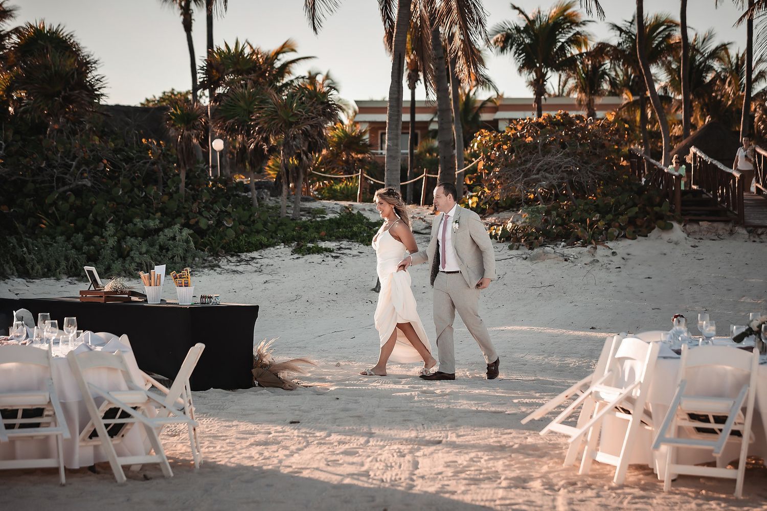 Bride and groom walking hand in hand on a sandy beach wedding venue with white chairs and palm trees at sunset.
