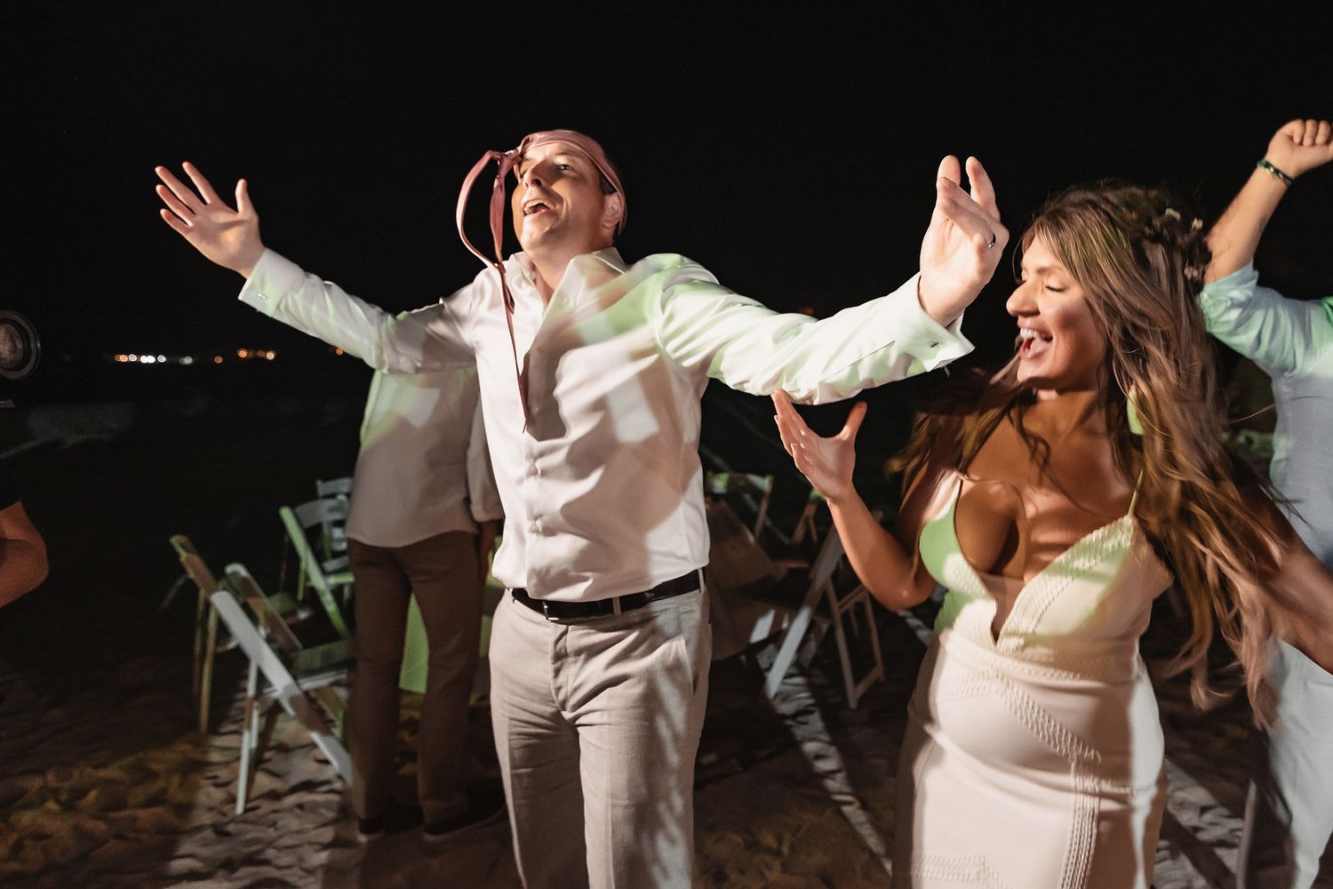 Joyful bride and groom dancing outdoors at night during wedding reception celebration on beach.