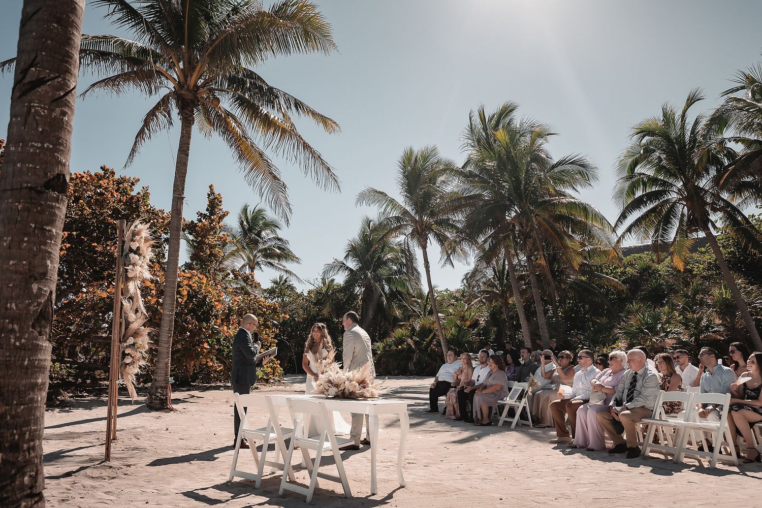 Outdoor beach wedding ceremony with guests seated on white chairs under tropical palm trees on a sunny day.