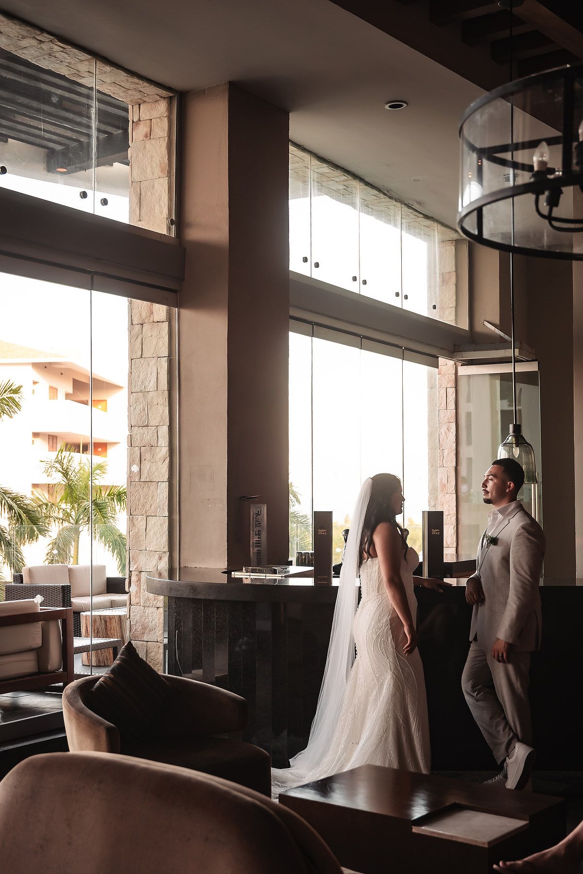 Bride and groom share a romantic first look moment in an elegant hotel lobby with large windows.