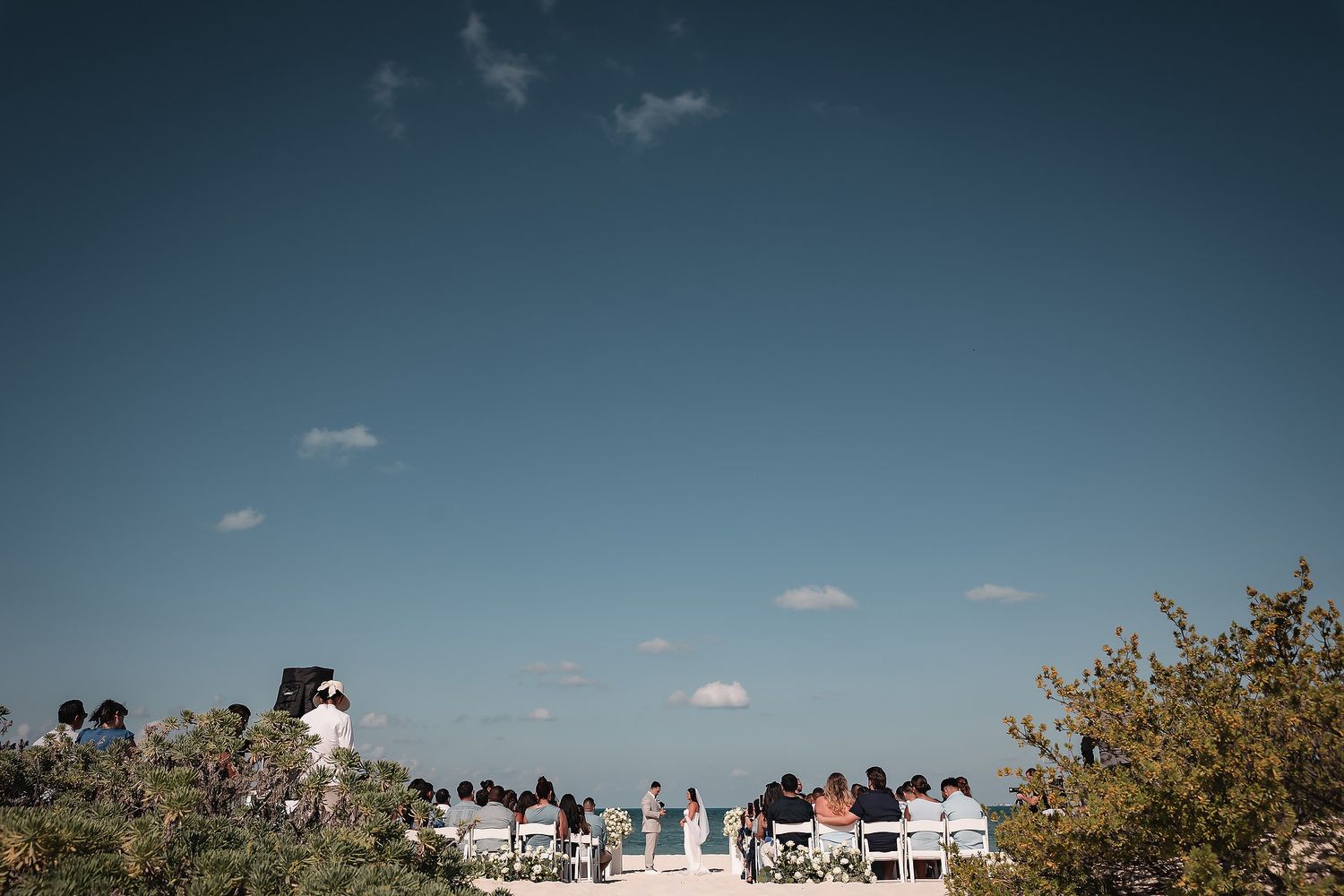 Outdoor wedding ceremony with guests seated in white chairs under a clear blue sky surrounded by lush greenery.