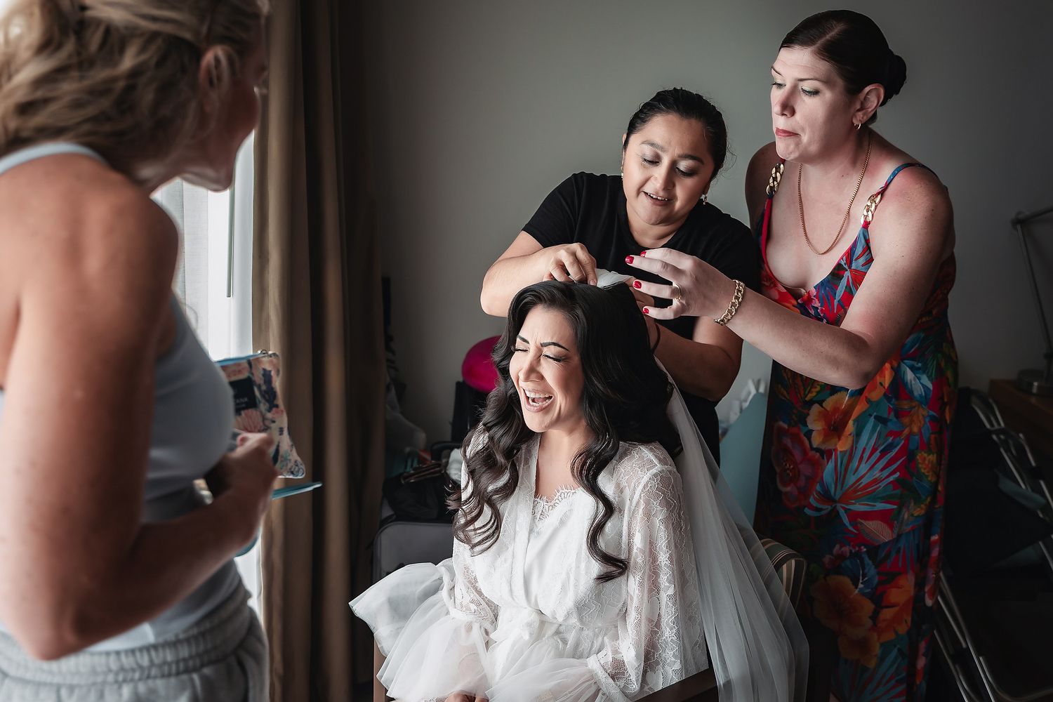 Bride smiling joyfully while two stylists arrange her hair before her wedding ceremony.