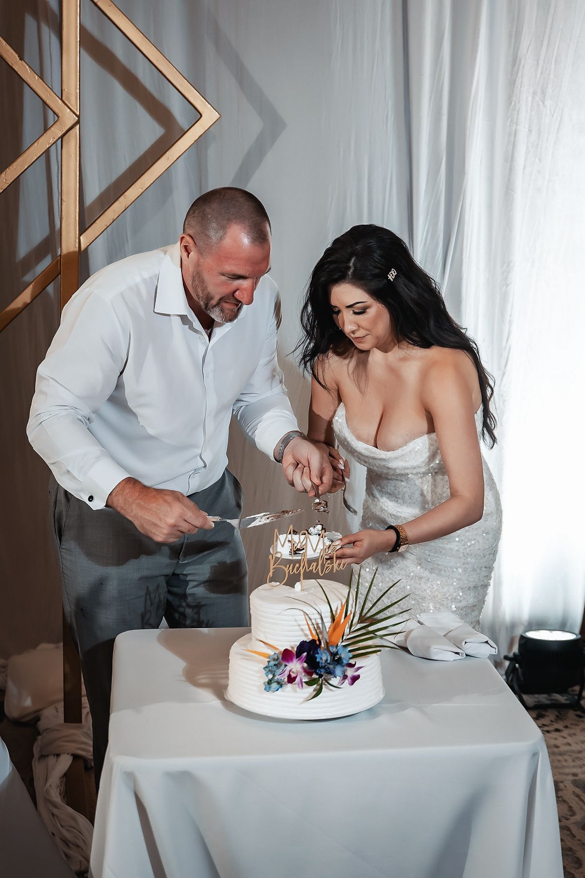 Bride and groom cutting white wedding cake decorated with tropical flowers at elegant reception venue.