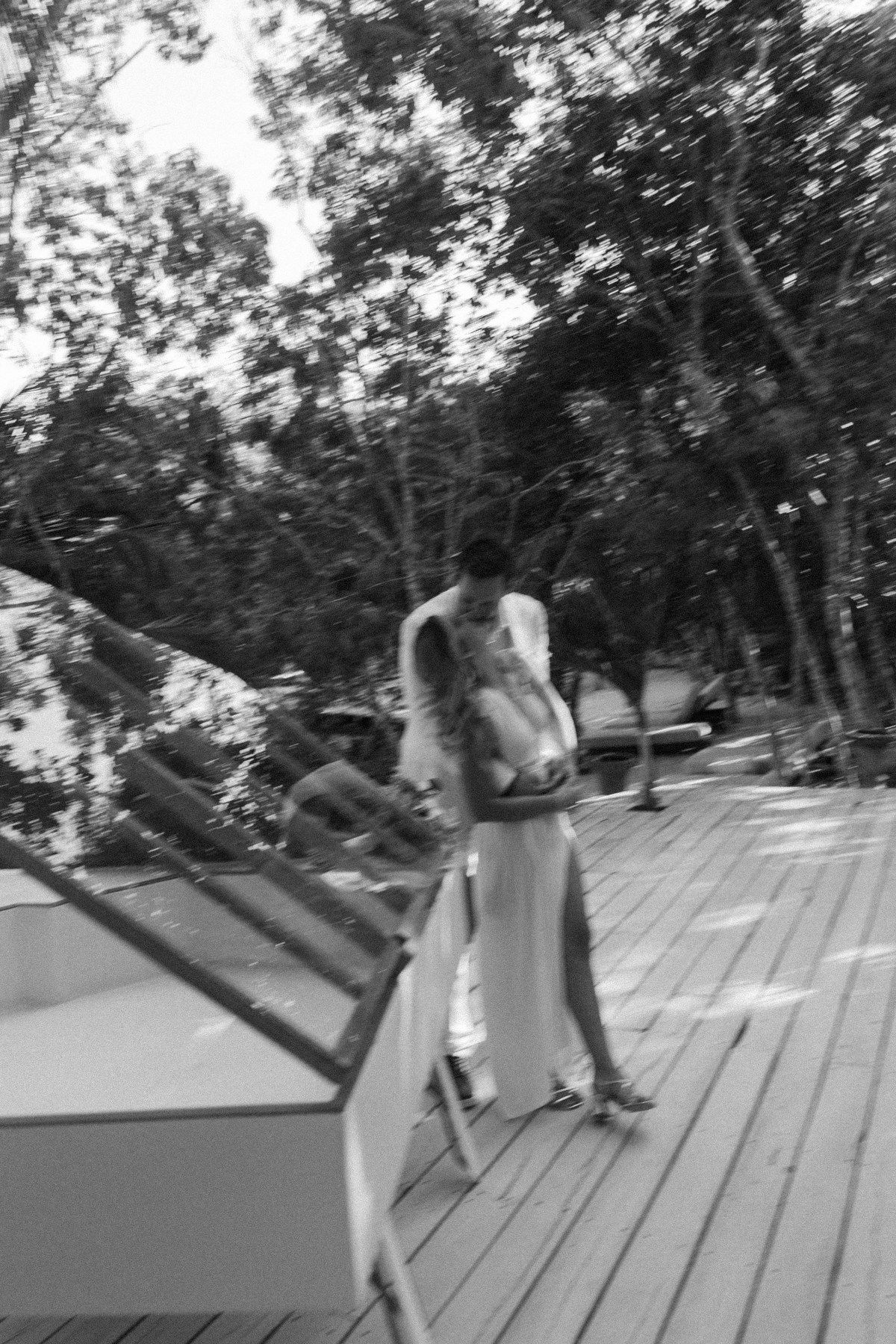 Black and white photo of a woman in a long dress walking on a wooden deck surrounded by lush trees.