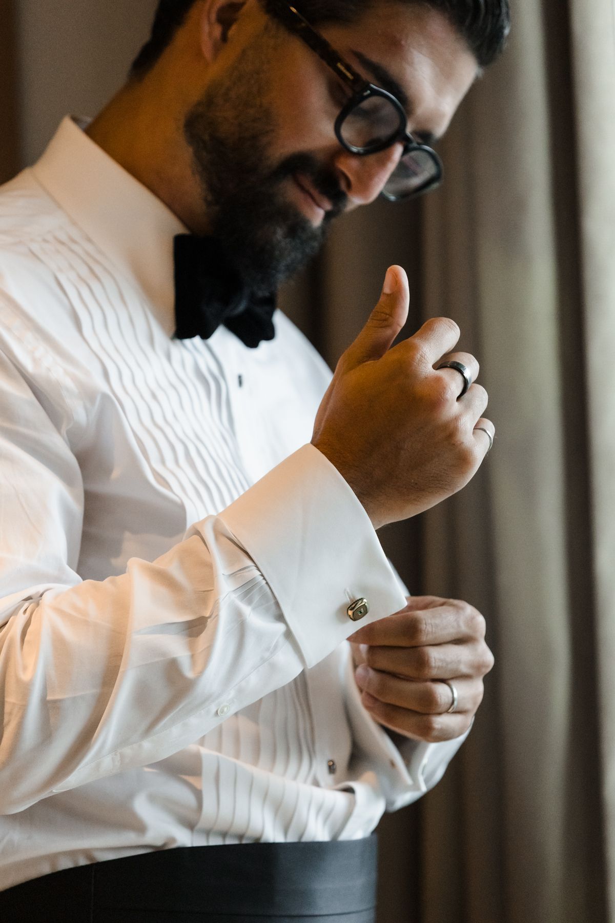 A bearded groom in a white tuxedo shirt and glasses fastens his gold cufflinks while smiling.