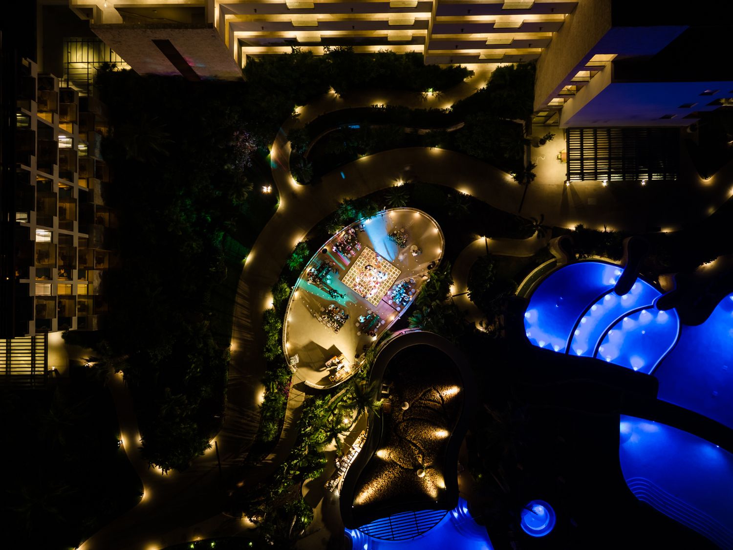 Aerial night view of a luxury hotel courtyard with glowing curved pathways and a blue illuminated pool.