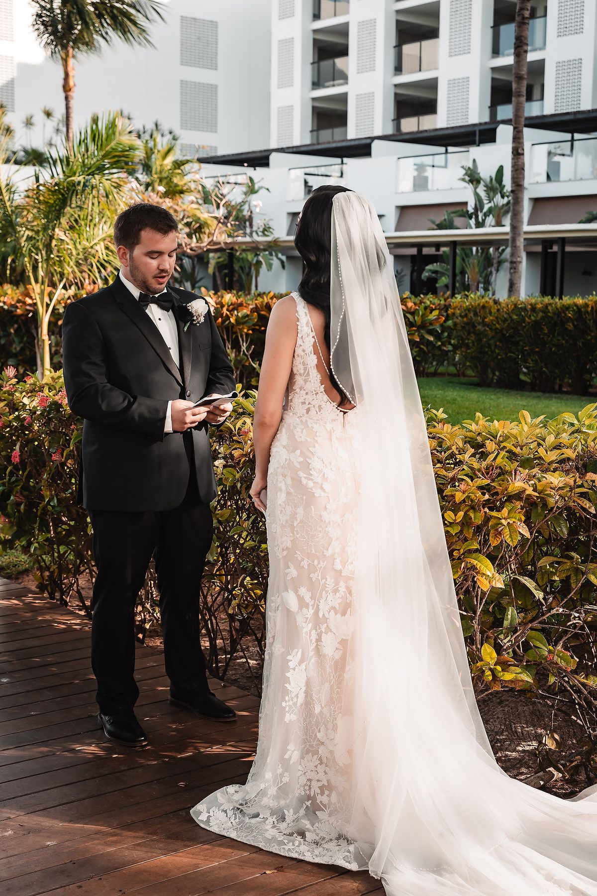 Groom reads vows from card to bride in lace wedding dress and veil outdoors at tropical resort wedding.
